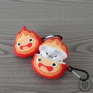 May include: A pair of red and orange silicone AirPod cases, each shaped like a cartoon fire demon, with black carabiner clips. One case is open, showing white AirPod earbuds. The design features a smiling face with black eyes and a red mouth.