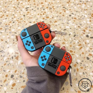 Op de afbeelding: Twee beschermhoezen voor draadloze oordopjes, ontworpen om op Nintendo Switch-controllers te lijken. Elke hoes is verdeeld in blauwe, rode en grijze secties, met knopdetails en de tekst "Nintendo Switch" op de voorkant. Inclusief een zilveren karabijnhaak.