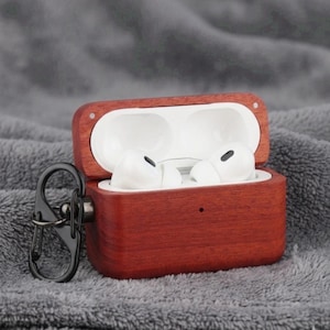 May include: A wooden AirPods case with a rich, reddish-brown finish, open to reveal white earbuds. The case has a black metal carabiner clip attached. The background is a soft, gray fabric.