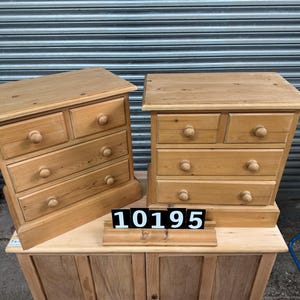 May include: Two small, light-coloured wooden bedside cabinets with four drawers each. The cabinets have rounded knobs and are displayed on a larger wooden cabinet. The number "10195" is displayed on a small stand.