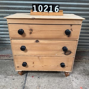 May include: A three-drawer wooden chest with black knobs. The chest is made of light-coloured wood and has a slightly angled design. The number "10216" is displayed on top. The chest is a piece of furniture.