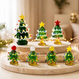May include: A collection of seven miniature glass Christmas trees in varying sizes. Each green tree features white glass garland, colorful glass ornaments, and a solid star or sphere topper. The trees are arranged on two tiered circular stone platforms.
