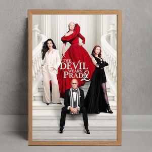 Könnte beinhalten: Ein gerahmtes Poster mit dem Filmtitel "Der Teufel trägt Prada 2" in einem hellen Holzrahmen. Das Poster zeigt die Charaktere in formeller Kleidung, wobei ein rotes Kleid im Mittelpunkt steht. Der Titel ist weiß.