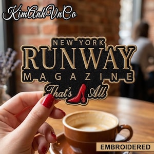 May include: Embroidered patch with the text "NEW YORK RUNWAY MAGAZINE That's All" in gold and black. A red high heel is featured in the design. The patch is held in front of a coffee cup.