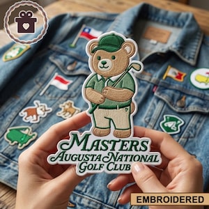 Op de afbeelding: Geborduurde patch met een beer in golfoutfit, met een club. De patch zegt "Masters Augusta National Golf Club" in groen schrift. De patch wordt voor een spijkerjasje met andere patches en vlaggen gehouden.