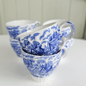 May include: A collection of white ceramic cups featuring a blue transferware pattern depicting a rural landscape with buildings, trees, and clouds. Each cup has a curved handle and a flared rim, showcasing a consistent blue and white design.