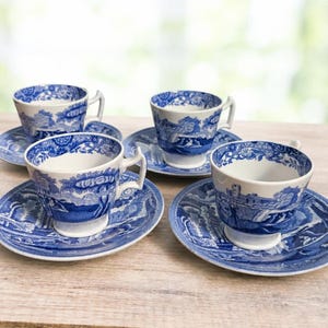May include: A set of four white ceramic teacups and matching saucers, each decorated with a detailed blue transferware pattern depicting traditional landscape scenes with trees, buildings, and floral borders on the rims and cup exteriors.