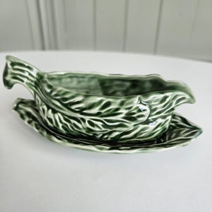 May include: A dark green ceramic gravy boat with an attached underplate, featuring a moulded leaf texture throughout the exterior. The piece has a glossy finish, a handle on one side, and a pouring spout on the opposite end, resembling a cabbage leaf.