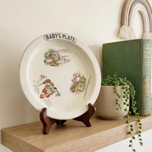 May include: A round, off-white enamel baby's plate featuring black text that reads BABY'S PLATE at the top. The plate is decorated with three colorful illustrations of nursery rhyme characters and text, supported by a dark brown wooden stand.