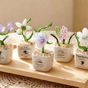 Puede incluir: Una colección de esculturas de flores de vidrio en pequeñas macetas blancas, cada una con un diseño floral único y tallos verdes. Las macetas tienen texto con temática de graduación. Las flores son en tonos de morado, blanco y rosa, sobre una bandeja de madera.