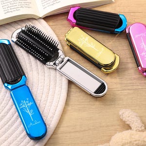 May include: A collection of colorful, compact hairbrushes. One blue brush is open, revealing a comb and mirror. Other brushes are closed, in pink, gold, and black. Each has a decorative design and text. The brushes are on a light-colored surface.