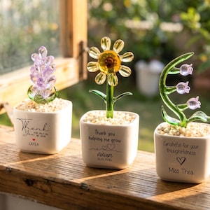 Peut inclure: Trois sculptures de fleurs en verre dans des pots carrés blancs. Les fleurs sont une branche de lavande, un tournesol jaune et une tige verte avec des fleurs roses en forme de cloche. Chaque pot a un message et un nom.