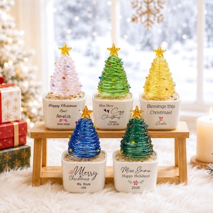 Peut inclure: Cinq sapins de Noël miniatures en verre dans des pots en céramique blanche, chacun surmonté d'une étoile jaune. Les arbres sont roses, verts, jaunes, bleus et vert foncé. Chaque pot porte un texte personnalisé, incluant les noms Amelia, Mary, Sadie, Blaik et Emma.
