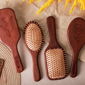 May include: Four wooden hairbrushes with different shapes and floral engravings. Two brushes have metal bristles. One brush is engraved with the name "Soren", and another with "Elowen". The brushes are arranged on a neutral-toned surface.