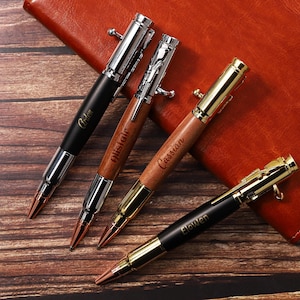 Op de afbeelding: Vier gepersonaliseerde pennen met kogelvormige ontwerpen. De pennen hebben zwarte, zilveren en gouden accenten, met houten en metalen vaten. Elke pen is gegraveerd met een naam. De pennen zijn gerangschikt op een houten oppervlak naast een leren notitieboek.