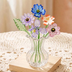 Puede incluir: Un jarrón de cristal transparente con un ramo de flores de cristal de colores. Las flores incluyen flores azules, moradas, rosas y naranjas con tallos y hojas verdes. El jarrón está sobre una base de madera.