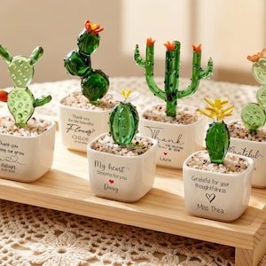 May include: Six miniature glass cactus figurines in white square ceramic pots filled with small pebbles. Each pot features black printed text and names including Thank you helping me grow, Flowers, My heart blooms for you, Thank, and Grateful.
