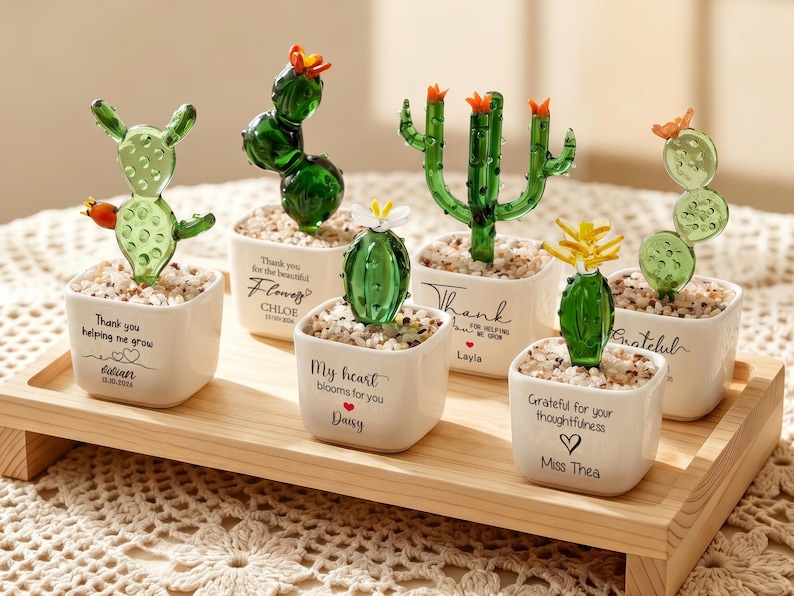 May include: Six miniature glass cactus figurines in white square ceramic pots filled with small pebbles. Each pot features black printed text and names including Thank you helping me grow, Flowers, My heart blooms for you, Thank, and Grateful.