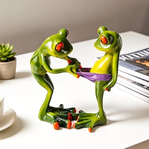 May include: A whimsical green frog figurine with red eyes and feet, holding a purple object. The frog has a yellow belly and is posed with another frog. The figurine is on a white surface with a cup of coffee and plants in the background.