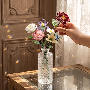 May include: A clear glass vase with a diamond pattern holds a colorful bouquet of glass flowers. The flowers include white, pink, purple, and red blooms with green leaves. The vase sits on a wooden table.