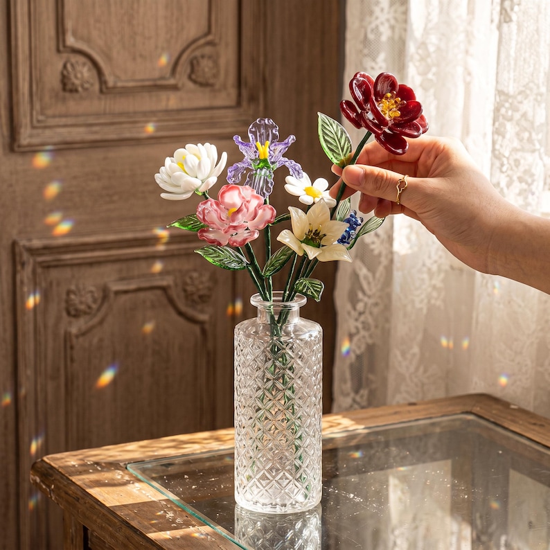 May include: A clear glass vase with a diamond pattern holds a colorful bouquet of glass flowers. The flowers include white, pink, purple, and red blooms with green leaves. The vase sits on a wooden table.