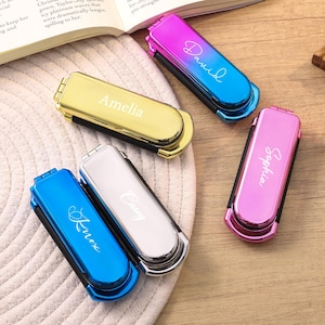 May include: A collection of personalized nail clippers in various colors, including gold, silver, blue, pink, and a gradient of blue to pink. Each clipper has a name engraved in a cursive font: Amelia, Casey, Knox, David, and Sophia. The clippers are on a wooden surface.