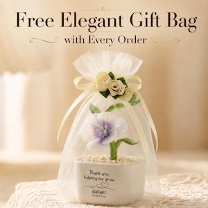 May include: A white ceramic planter containing a small artificial flower with purple and white petals, green leaves, and a base of light-colored pebbles. The planter is printed with the text Thank you helping me grow, Vivian, and 13.10.2026.