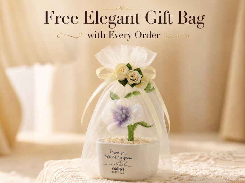 May include: A white ceramic planter containing a small artificial flower with purple and white petals, green leaves, and a base of light-colored pebbles. The planter is printed with the text Thank you helping me grow, Vivian, and 13.10.2026.