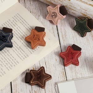Puede incluir: Un conjunto de marcadores de cuero en forma de estrella en varios colores, incluyendo marrón, rosa, verde y rojo. Cada marcador presenta iniciales personalizadas o la palabra "DAD" y un diseño decorativo. Tienen una banda elástica marrón.