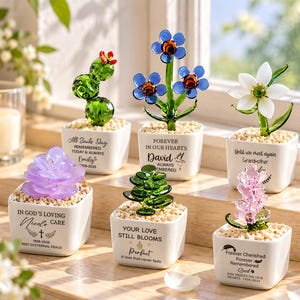 May include: Six glass plant figurines in white square pots filled with small stones. Each pot features unique memorial text and names. The plants include a green cactus, blue flowers, a white flower, a purple succulent, a green succulent, and a pink flower.