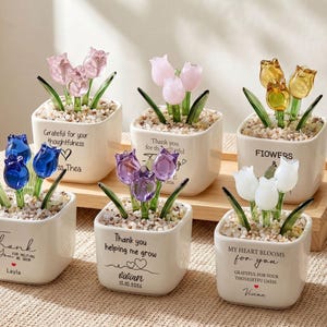 Peut inclure: Six pots carrés en céramique blanche contenant des tulipes en verre rose, bleu, violet, jaune et blanc. Chaque pot présente des feuilles en verre vert, des petits cailloux et du texte imprimé incluant des messages de remerciement et des prénoms.