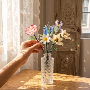 May include: A clear glass vase with a diamond pattern holds a bouquet of colorful glass flowers. The flowers include a pink rose, blue hyacinths, white daisies, and cream-colored lilies. The vase sits on a wooden table near a window.