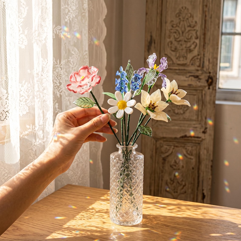 May include: A clear glass vase with a diamond pattern holds a bouquet of colorful glass flowers. The flowers include a pink rose, blue hyacinths, white daisies, and cream-colored lilies. The vase sits on a wooden table near a window.