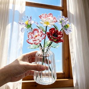 May include: A clear glass vase with a ribbed design holds a bouquet of colorful glass flowers. The flowers include red, white, and purple blooms with green leaves. The vase is held in front of a window with a blue sky visible.
