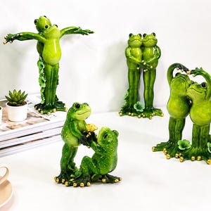Könnte beinhalten: Eine Sammlung grüner Froschfiguren in verschiedenen Posen. Ein Frosch hat die Arme ausgestreckt, während andere sich umarmen oder Händchen halten. Die Frösche haben goldene Akzente und stehen auf einer weißen Oberfläche. Kleine Sukkulenten in Töpfen und eine Tasse Kaffee sind ebenfalls vorhanden.