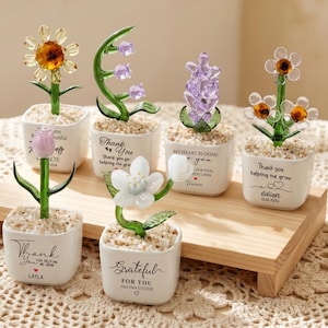 Puede incluir: Una colección de esculturas de flores de vidrio en pequeñas macetas blancas, cada una con un diseño y color únicos. Las macetas tienen mensajes como "Gracias" y "Agradecido por ti". Las flores se exhiben en una bandeja de madera.