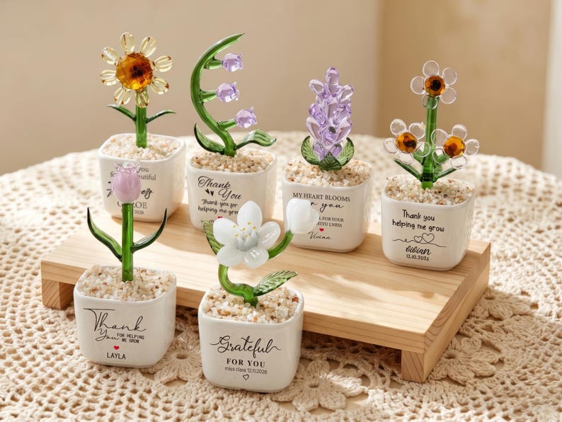 May include: Six glass flower figurines in small white square ceramic pots filled with pebbles. Each pot features black printed text including phrases like Thank You, Grateful For You, and My Heart Blooms, along with personalized names and dates.