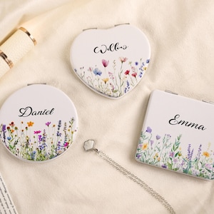 May include: Three personalized compact mirrors in different shapes: round, heart, and square. Each white mirror features a floral design with colorful wildflowers and a name in elegant script: Daniel, Willow, and Emma. A pen and necklace are also visible.
