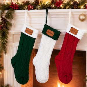 May include: Three cable-knit Christmas stockings hanging in a row. The left is dark green with a white cuff, the center is white with a dark green cuff, and the right is red with a white cuff. Each features a brown leather tag with a name.