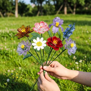 May include: A bouquet of colorful glass flowers, including a daisy, iris, and rose, held in a person's hands. The flowers are in shades of red, pink, purple, yellow, and brown, with green stems and leaves. The background is a blurred green field.