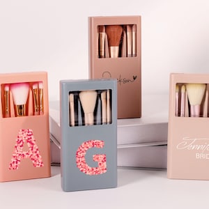 Op de afbeelding: Vier make-up kwastensets in verschillende gekleurde cases. De cases zijn roze, grijs en bruin. Elke case heeft een andere initiaal of naam erop. De kwasten zijn zichtbaar door een rechthoekig venster aan de voorkant.