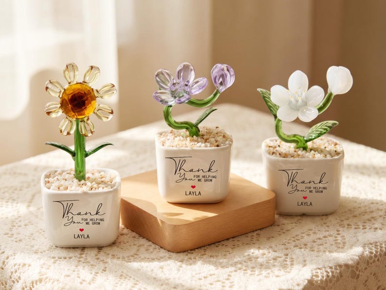 May include: Three glass flowers in white ceramic pots filled with small stones. Each pot features the black text Thank You for helping me grow and the name LAYLA with a red heart. The flowers are yellow, purple, and white with green stems.