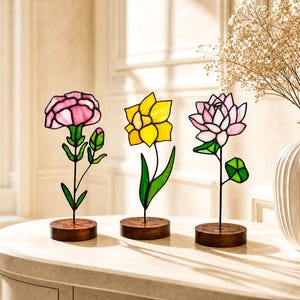 以下が含まれることがあります： 円形の木製台座に飾られた3つのステンドグラス製の花の彫刻。左はピンクのカーネーション、中央は黄色の水仙、右はピンクと白の蓮の花です。それぞれの花には緑色のガラスの葉と細い金属製の茎がついています。