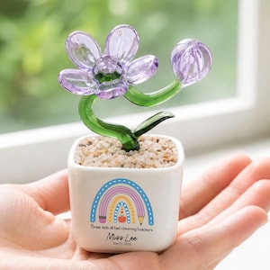 May include: A small white square ceramic planter filled with light-colored pebbles and a glass flower sculpture with purple petals and a green stem. The planter features a colorful rainbow graphic, a pencil, an apple, and the text "Brave kids all had cheering teachers Miss Lee June 15, 2026."