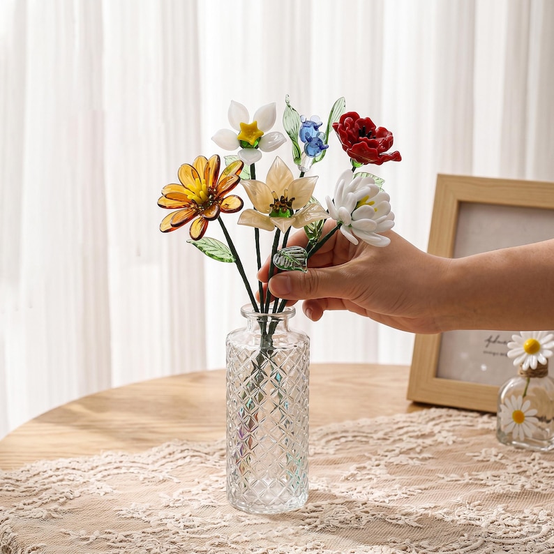 May include: A clear glass vase with a diamond pattern holds a bouquet of colorful glass flowers. The flowers include yellow, white, red, and blue blooms with green stems and leaves. The vase is on a wooden table.