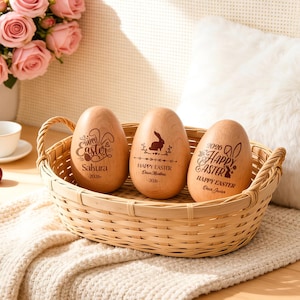 May include: Three wooden Easter eggs with engraved "Happy Easter" text and bunny designs sit in a woven basket. The eggs are light brown, and the basket is a natural tan color. A bouquet of pink roses and a white pillow are in the background.