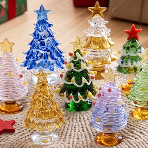 May include: A collection of eight miniature glass Christmas tree figurines in various colors including blue, gold, green, pink, and light purple. Each tree features a star topper and decorative glass accents, standing on a circular base.