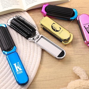 May include: Several personalised folding hairbrushes in blue, gold, pink, and black. Each brush has a mirror and is engraved with a name or initial. The blue brush displays "K Knox".