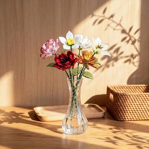 May include: A clear glass vase with a spiral design holds a colorful bouquet of artificial flowers. The arrangement includes pink, red, white, and orange blooms with green leaves. The vase sits on a wooden surface, with a woven basket and a wooden tray in the background.
