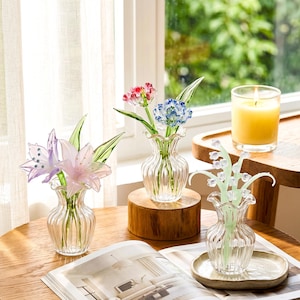 Pode incluir: Vasos de vidro transparente com flores de vidro coloridas, incluindo flores rosa, roxas, vermelhas e azuis. Os vasos estão sobre uma mesa de madeira com uma vela acesa e uma revista.